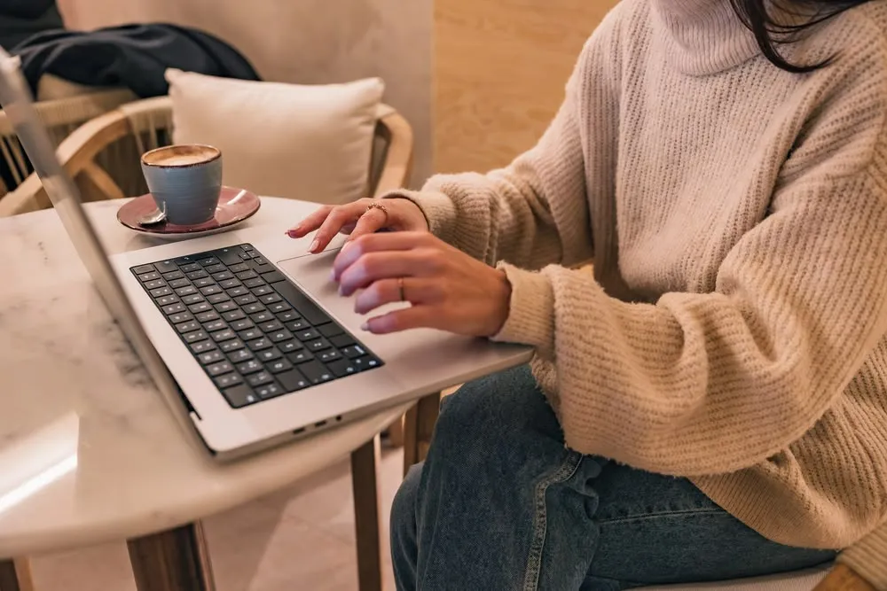 A person in a cream knit sweater typing on a laptop at a marble cafe table beside a cup of coffee